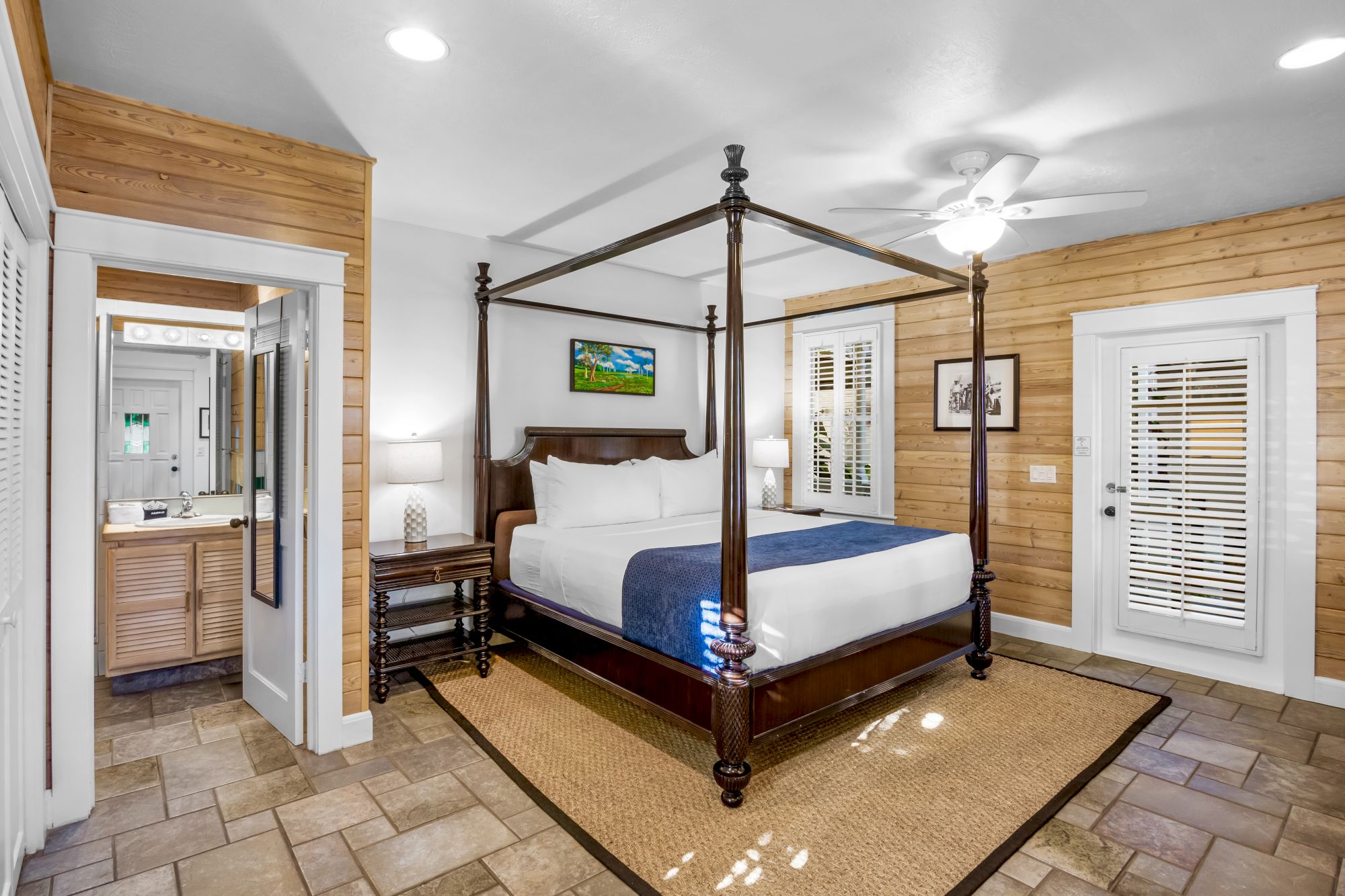 This image shows a cozy bedroom with a four-poster bed, wooden walls, a ceiling fan, a bathroom entrance, and a door with blinds.