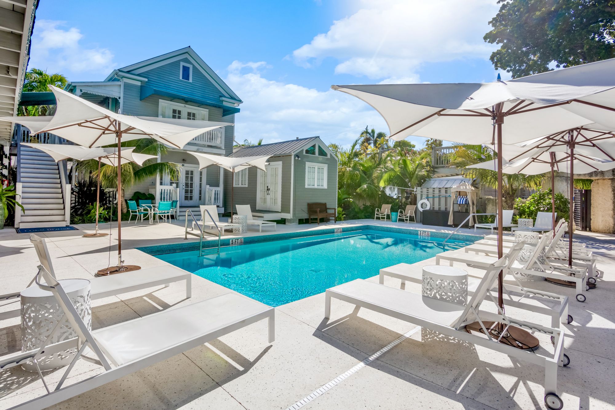 A sunny pool area with white lounge chairs and umbrellas, surrounded by a two-story house and lush greenery, under a blue sky.