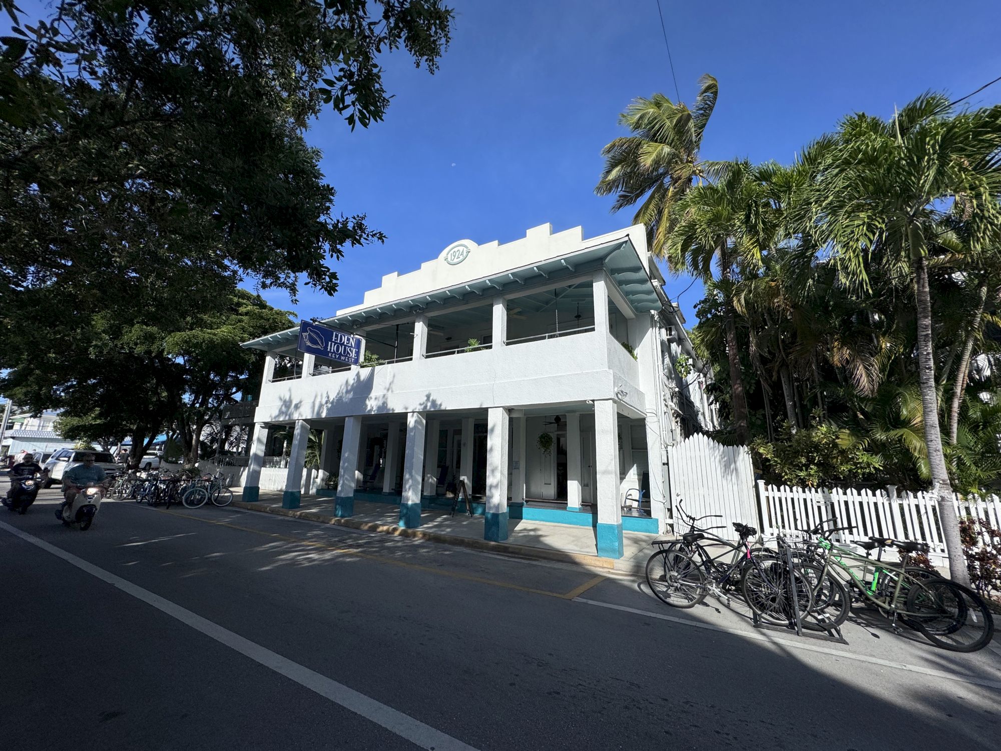 The image shows a white two-story building with bicycles parked outside. Palm trees surround it under a clear blue sky.