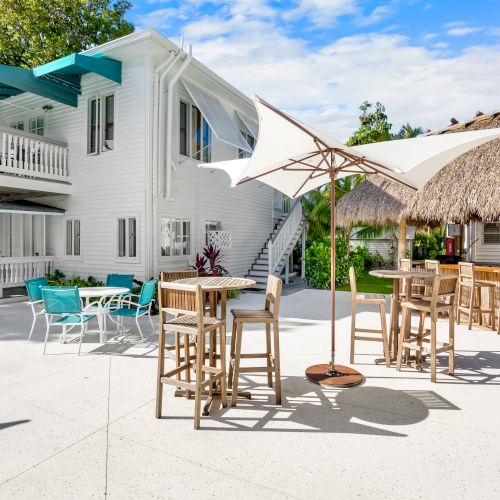 The image shows an outdoor patio with tables, umbrellas, and chairs, next to a two-story white building and a thatched-roof bar area.