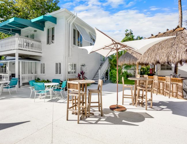 The image shows an outdoor patio with tables, umbrellas, and chairs, next to a two-story white building and a thatched-roof bar area.