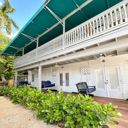 The image shows a two-story white building with a green awning, featuring outdoor seating and surrounded by greenery and palm trees.