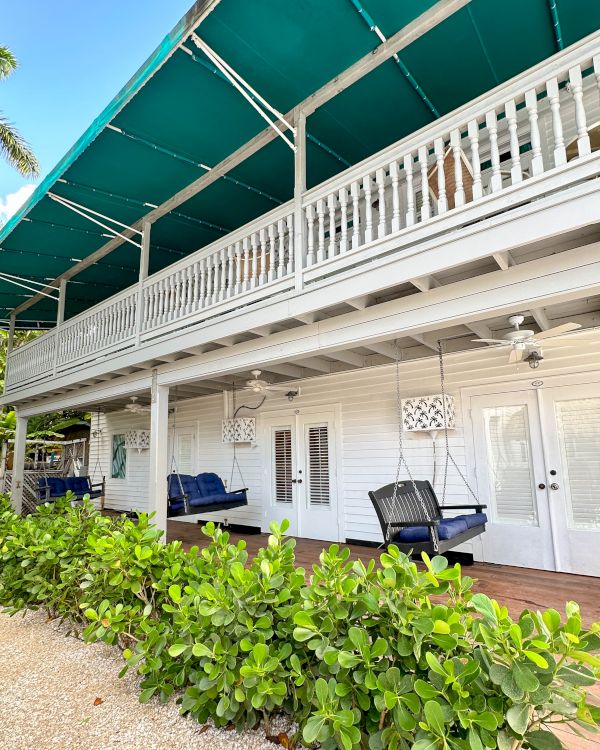 The image shows a two-story white building with a green awning, featuring outdoor seating and surrounded by greenery and palm trees.