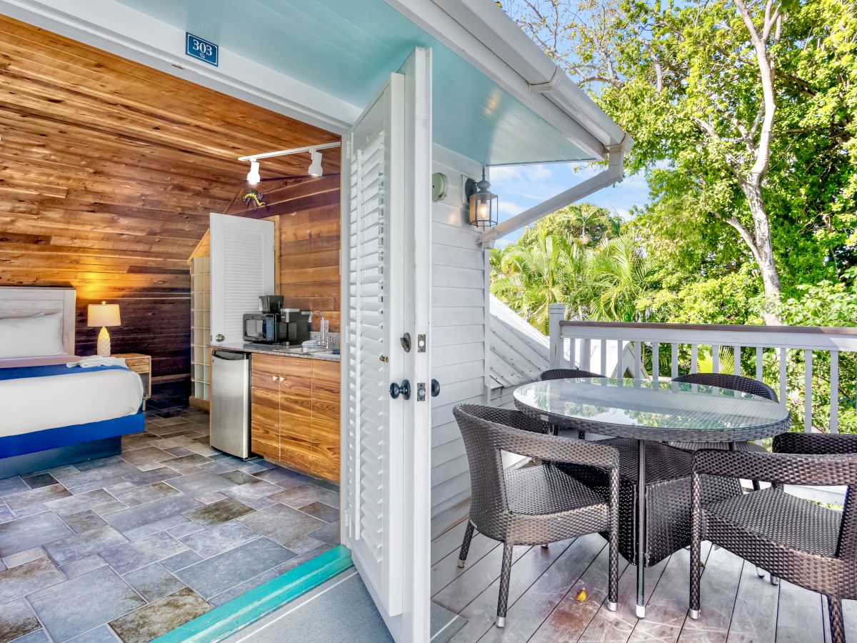 A cozy room with a bed and a kitchenette opens to a deck with a wicker table set, surrounded by lush greenery outside.