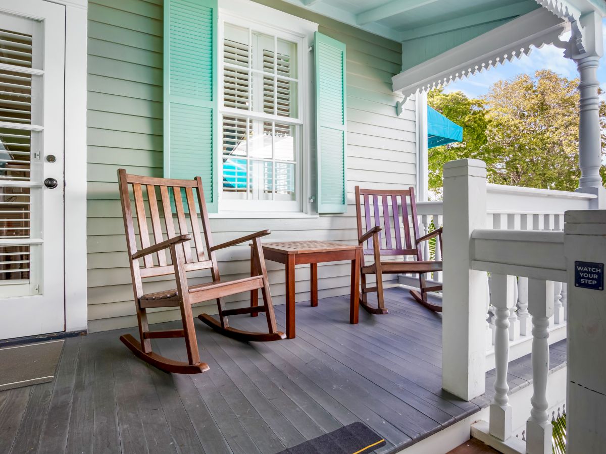 A cozy porch with two wooden rocking chairs, a small table, and pastel-colored shutters, overlooking a lush garden.