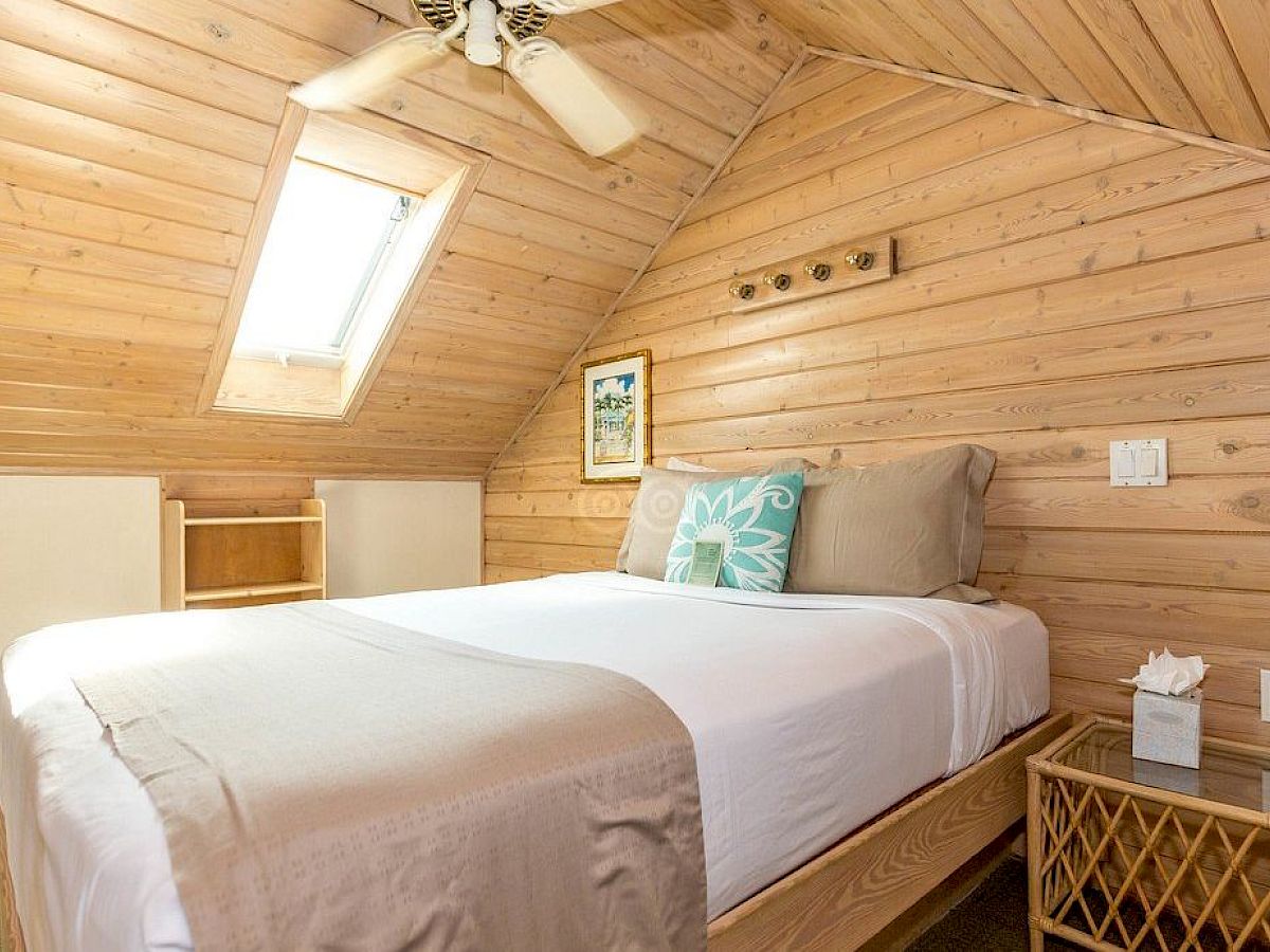 A cozy attic bedroom with wood paneling, a skylight, a neatly made bed, a side table, and a small shelf.