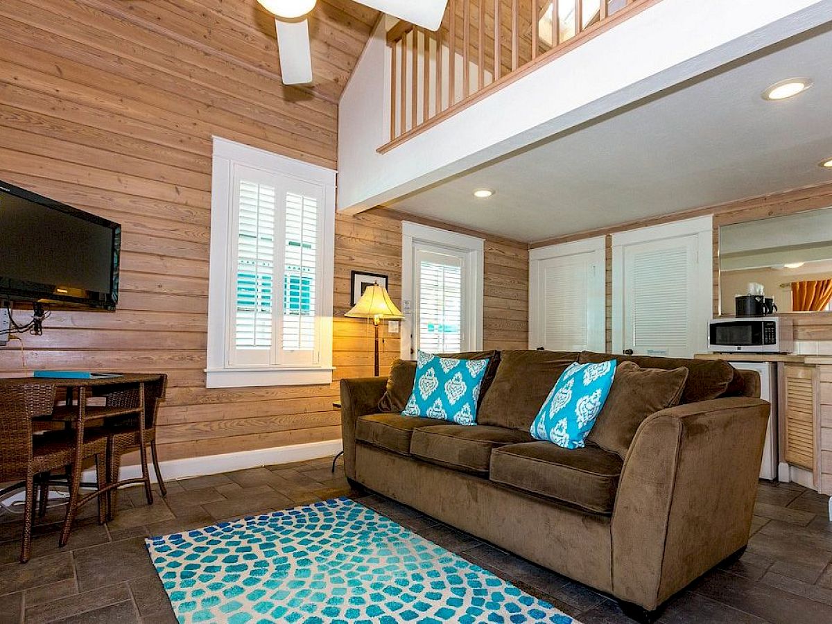 A cozy living room with a sofa, patterned cushions, a TV, small dining area, and kitchen, featuring wood paneling and modern decor.