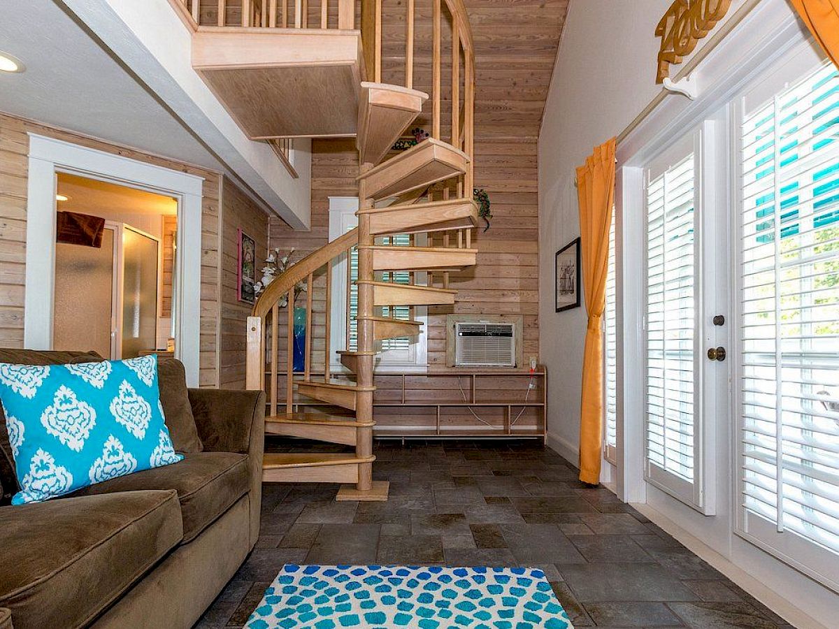 A cozy room with a spiral staircase, brown sofa, patterned cushions, and a rug. Sunlight streams through large windows with curtains.