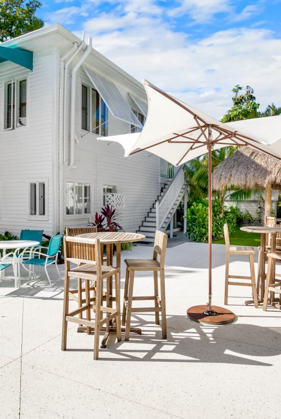 Outdoor area with a white house, patio furniture, umbrellas, and a tiki bar under a thatched roof on a sunny day.