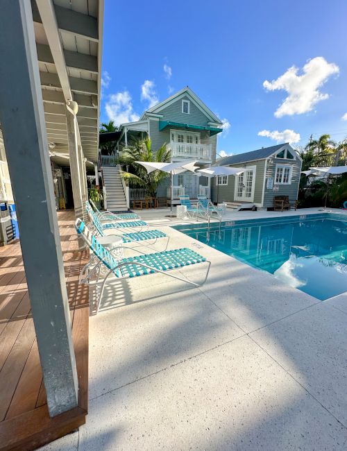 A sunny poolside area with lounge chairs, surrounded by a blue house and lush greenery. The sky is clear with a few clouds visible.