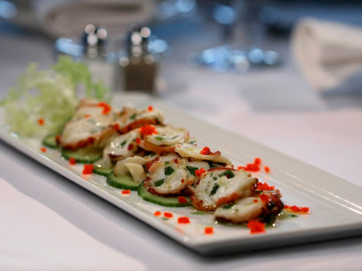 A plate of sliced octopus garnished with herbs and diced red peppers, served on cucumber slices with lettuce.