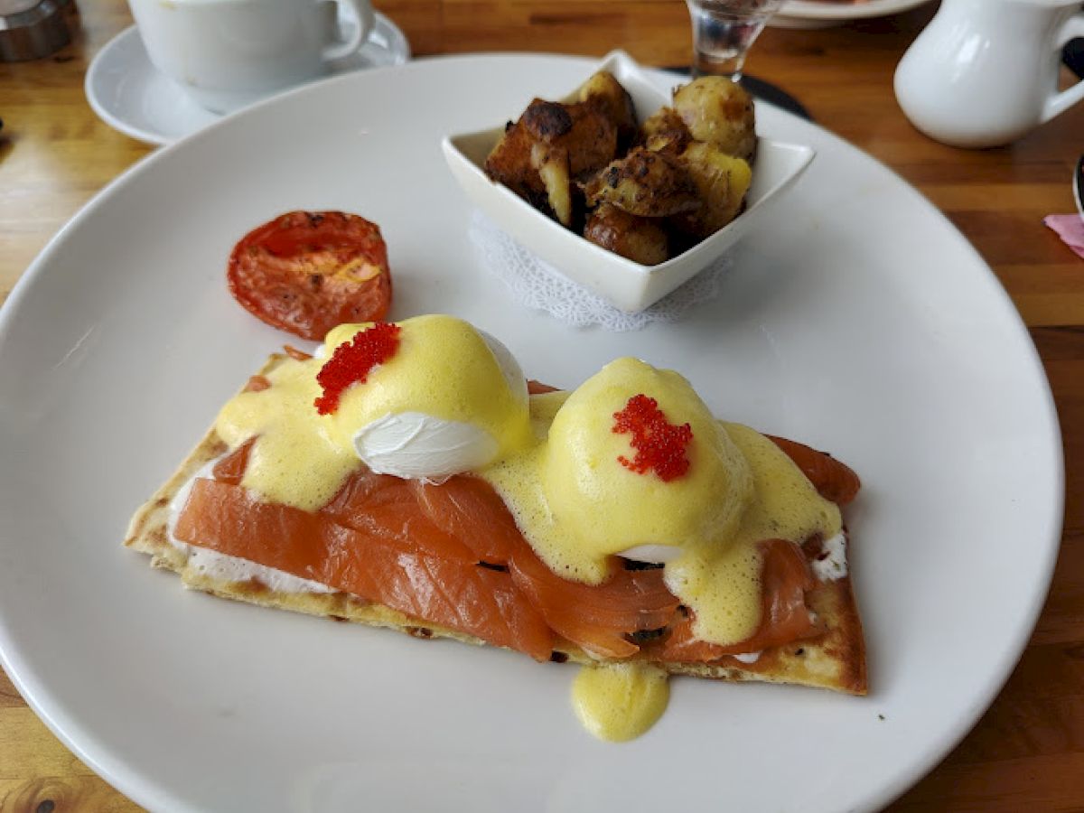 Eggs Benedict with smoked salmon on flatbread, topped with hollandaise and roe, served with roasted potatoes and a slice of grilled tomato.