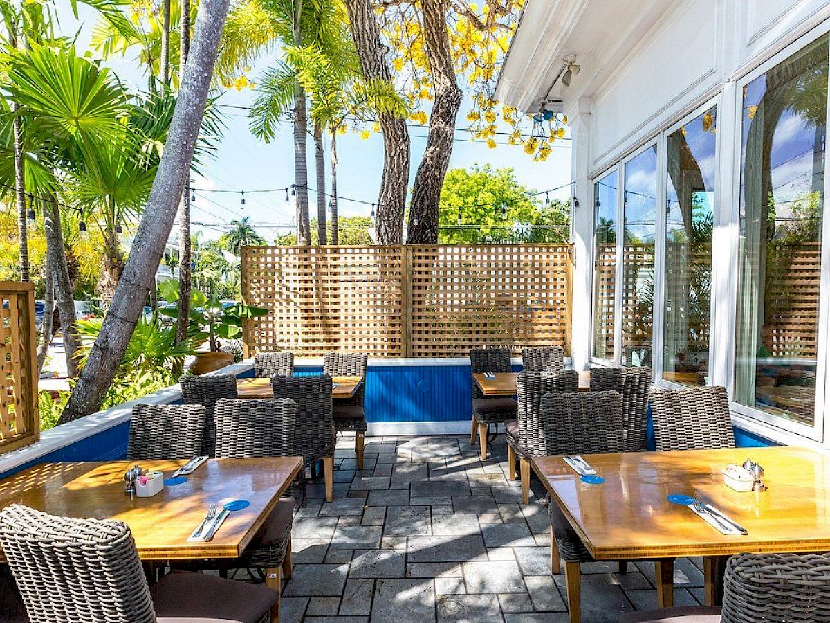 An outdoor patio with wicker furniture, wooden tables, lush greenery, and decorative lattices. Sunny ambiance and casual dining setup.