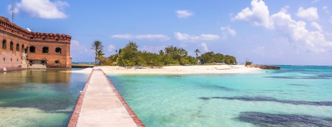 A walkway leads to a sandy island with lush greenery, surrounded by clear turquoise water under a blue sky with fluffy clouds.