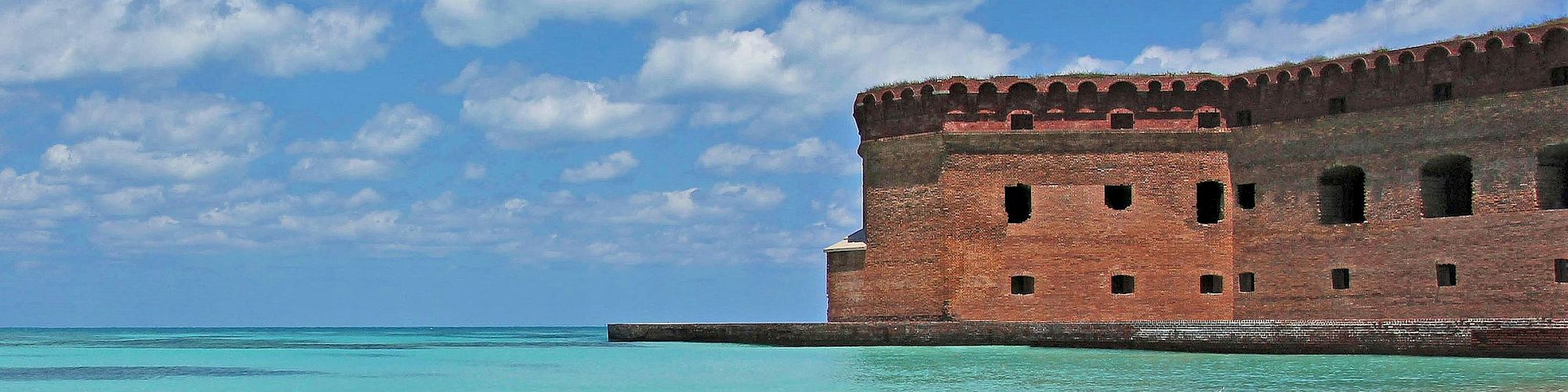 A sandy beach with clear turquoise water, a conch shell, and a historic brick fort along the shoreline under a blue sky with clouds.