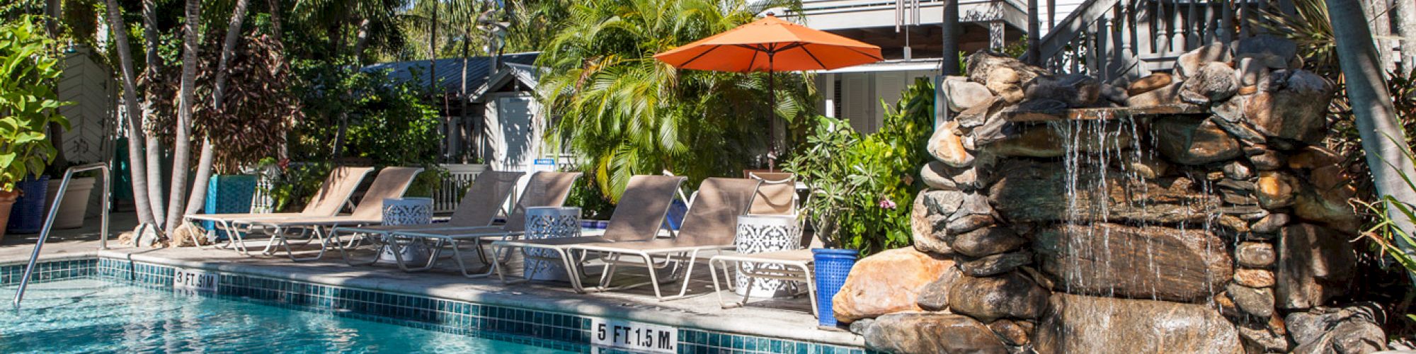 The image shows a tropical pool area with lounge chairs, an orange umbrella, palm trees, and a cascading rock waterfall feature.