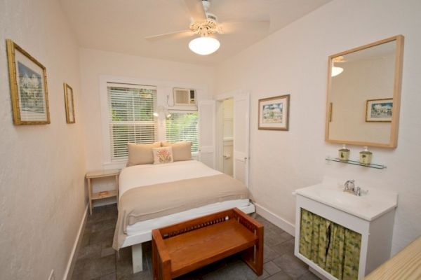 A cozy bedroom with a bed, wall art, a ceiling fan, a small dresser with a mirror, and a window. There's a small bench at the bed's foot.