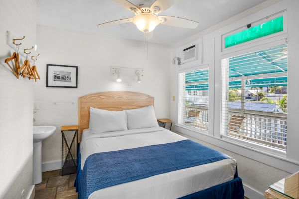 A small, bright bedroom with a double bed, blue blanket, wall-mounted hangers, sink, fan, and windows with green shades.