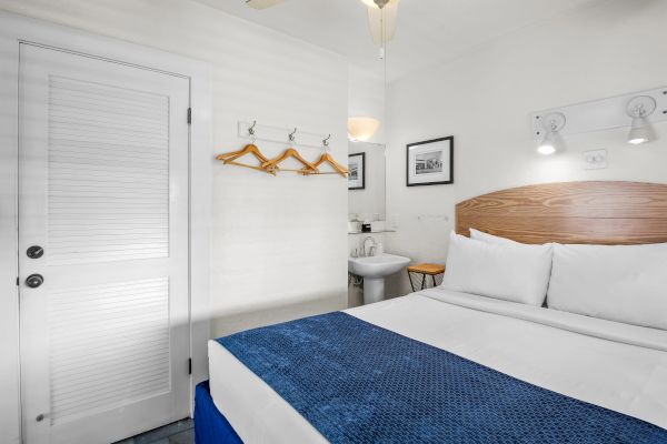 A small hotel room with a bed, blue blanket, two framed pictures, hooks with hangers, and a bathroom sink visible in the corner.