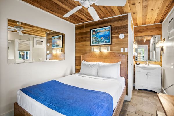 This image shows a cozy bedroom with a wooden ceiling and wall, a bed with a blue blanket, a mirror, artwork, and a sink area.