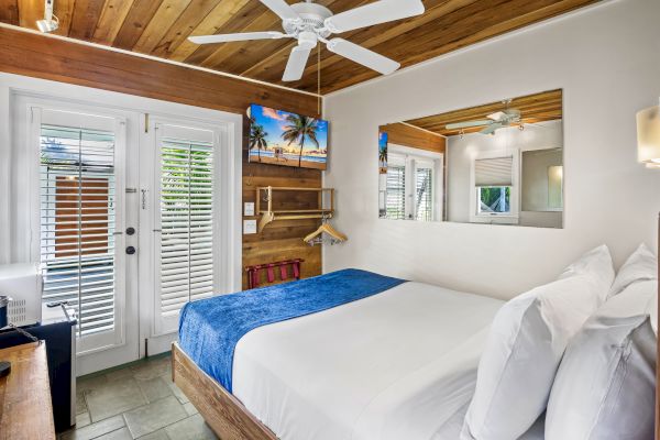 The image shows a cozy bedroom with a double bed, blue blanket, wooden accents, ceiling fan, TV, and a glass door with shutters leading outside.