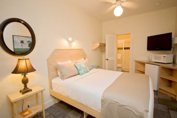 A cozy hotel room with a double bed, bedside lamp, wall mirror, TV, mini fridge, and bathroom. Decor is simple with a beach vibe.