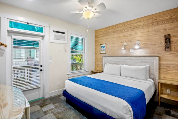 A cozy bedroom with a large bed, blue blanket, two nightstands, window, door, and ceiling fan, featuring wood accent wall and tile floor.