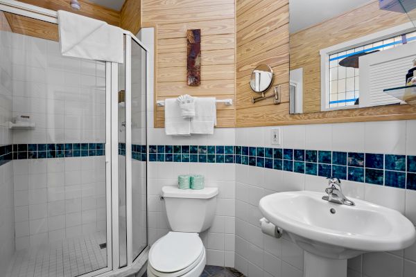 The image shows a small bathroom with a shower, sink, mirror, and toilet. Decorative tile accents and wooden panels are on the walls.