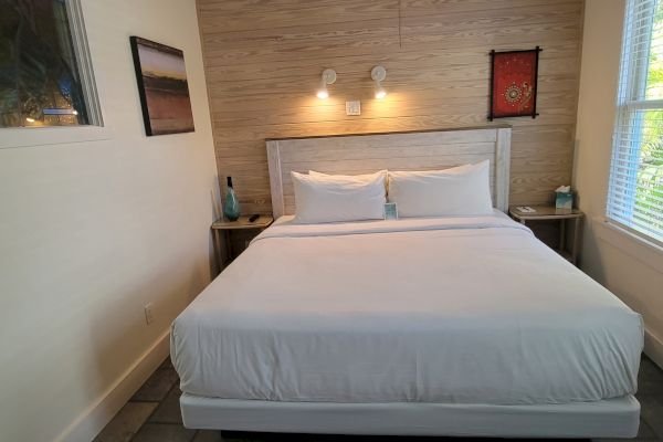 The image shows a neatly made bed with white linens in a cozy room, featuring wall art, bedside tables, and a window with blinds.