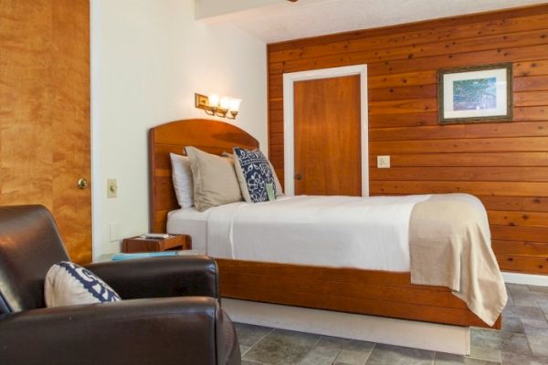 Cozy bedroom with wood paneling, bed with cushions, armchair, side table, and artwork on the wall, creating a warm and inviting space.