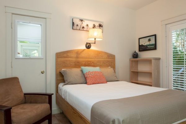 A cozy bedroom with a bed, pillows, chair, small shelf, wall art, and a lamp above the headboard. A window with blinds is to the right.