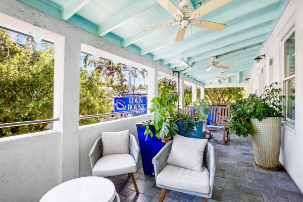 A porch with plants, comfy chairs, a ceiling fan, and a "Eden House" sign, surrounded by greenery and a relaxing atmosphere.