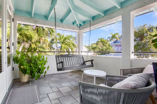 A cozy porch with a swing, wicker chairs, a small table, and lush plants. The ceiling is painted light blue, offering a tropical feel.