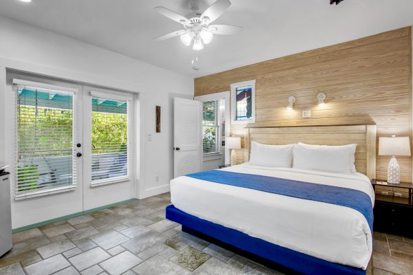 A modern bedroom with a king-sized bed, blue accents, wood paneling, two lamps, a ceiling fan, and glass doors leading outside.