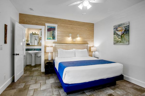 A modern bedroom with a large bed, blue bedspread, wall art, ceiling fan, and an open door leading to a bathroom with a sink and mirror.