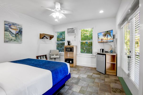 A bright room with a bed, TV, mini fridge, microwave, and decor. Features tiles, fan, and window shades. Tropical image on TV screen.