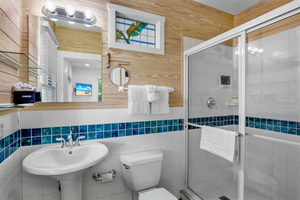 A modern bathroom with a glass shower, sink, toilet, and wooden accents. Blue tile detailing and a mirror above the sink complete the look.