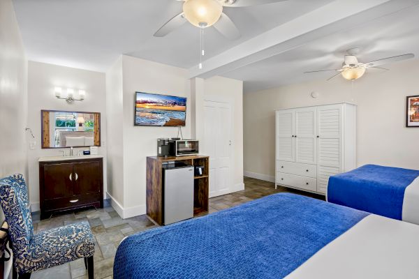 The image shows a hotel room with two beds, blue bedding, a wardrobe, a mini fridge, a microwave, a wall-mounted TV, and a small seating area.