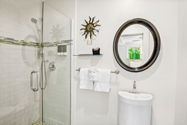 A bathroom with glass shower, round mirror, small sink, towels, and decorative wall piece.