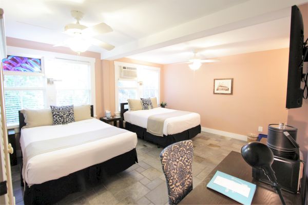 The image shows a hotel room with two beds, a desk, chair, TV, and ceiling fans. The room is well-lit with natural light from windows.