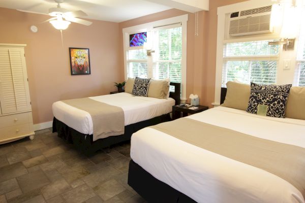 A cozy bedroom with two neatly made beds, decorative pillows, wall art, and a window. A ceiling fan and air conditioner are also visible.