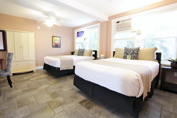 The image shows a tidy bedroom with two double beds, patterned cushions, a wardrobe, and a flat-screen TV on a pastel-colored wall.