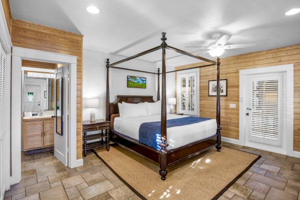 A cozy bedroom with a four-poster bed, blue bedding, wooden walls, a ceiling fan, and a doorway leading to a bathroom.