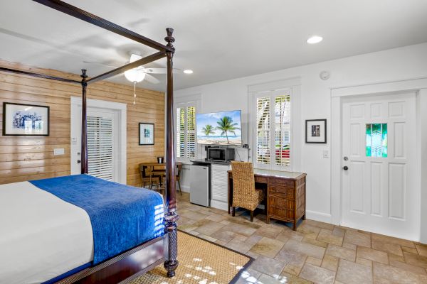 A cozy bedroom with a four-poster bed, desk, chair, microwave, and a TV displaying a beach scene. Light wood walls and tile flooring complete it.