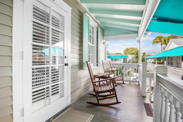 The image shows a porch with two wooden rocking chairs, white railings, French doors, and teal accents. It's a bright, relaxing setting.