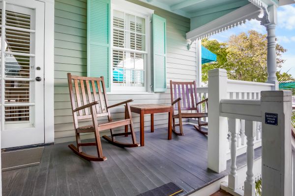 A cozy porch with two wooden rocking chairs, a small table, and green shutters. Surrounded by a peaceful outdoor setting.