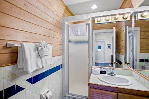 A bathroom with wood-paneled walls, a shower, a sink, and a mirror. Towels hang next to the shower along with a wall-mounted light fixture.