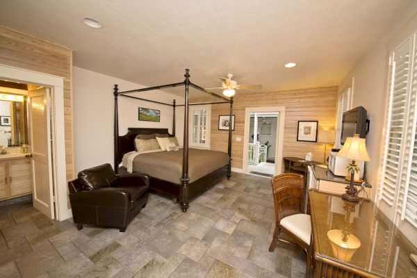 A well-furnished bedroom with a four-poster bed, armchair, desk, TV, and a connected bathroom. Walls are wood-paneled, flooring is tiled.