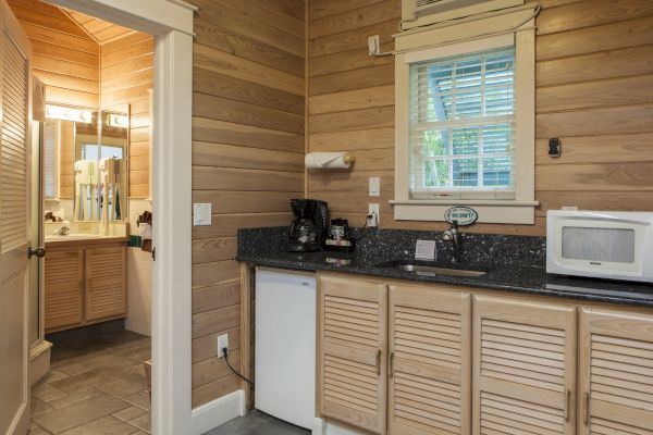 The image shows a small kitchenette with a fridge, microwave, and coffee maker, adjacent to a bathroom with a sink and wooden cabinets.