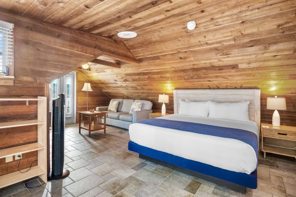 The image shows a cozy attic room with a bed, a couch, wooden walls and ceiling, a stand fan, and two bedside lamps for lighting.